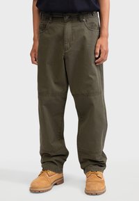 Pantalons cargo vert olive en tissu durable, avec une coupe droite, des poches latérales et des accents cousus aux genoux, associés à des chaussures beiges.
