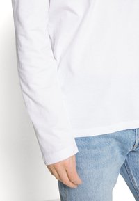 Weißes Langarm-Hemd aus Baumwolle mit lockerer Passform und sichtbaren Nähten, kombiniert mit hellblauen Jeans.