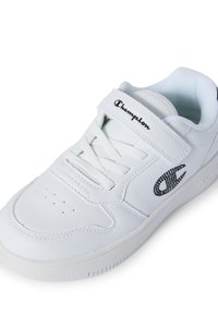 Sneaker bianco in materiale sintetico, con punta perforata, chiusura a laccetto e strap in velcro. Presenta un logo nero con strass. Suola in gomma flessibile.