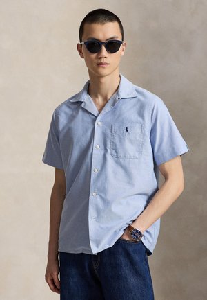 Homme portant des lunettes de soleil bleu marine, une chemise bleu clair à manches courtes avec une poche poitrine, un jean foncé et une montre marron avec un cadran bleu.