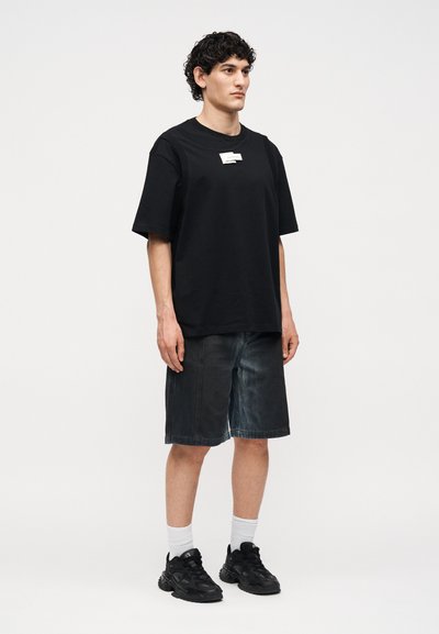 Νεαρός με σγουρά μαλλιά φοράει μαύρο φαρδύ T-shirt, σκούρα φαρδιά τζιν σορτς, λευκές κάλτσες και μαύρα chunky sneakers, στέκεται μπροστά σε λευκό φόντο.