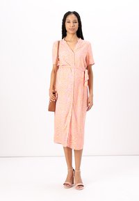 Pink dress with coral abstract pattern, short sleeves, and waist tie, paired with brown handbag and nude block heel sandals. Smooth fabric texture.