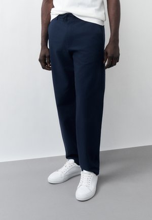 Άνδρας που φοράει μπλε σκούρο παντελόνι με κορδόνι, λευκά αθλητικά παπούτσια και λευκό αμάνικο τοπ μπροστά σε απλό ανοιχτό φόντο.