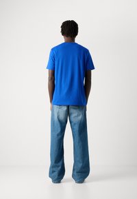 Persona con piel oscura y cabello rizado corto se encuentra frente a una pared blanca, vistiendo una camiseta azul brillante y pantalones vaqueros azules holgados.