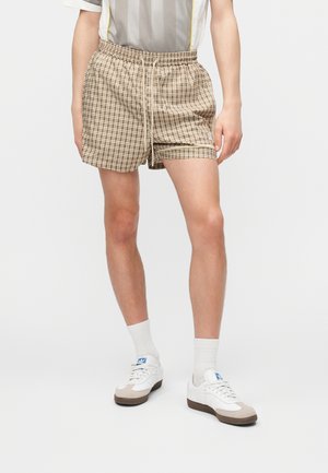 Άνδρας που φοράει μπεζ καρό σορτς, λευκά αθλητικά παπούτσια και λευκές κάλτσες πλήρους μήκους, στέκεται πάνω σε απλό φωτεινό φόντο.