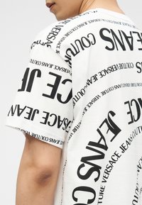 Μερική προβολή ενός ατόμου που φοράει λευκό t-shirt με μαύρο κείμενο "Versace Jeans Couture" τυπωμένο επανειλημμένα σε διάφορα μεγέθη και προσανατολισμούς.