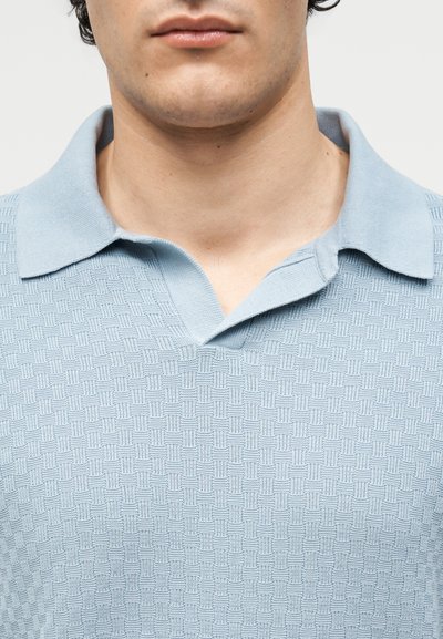 Κοντινό πλάνο ενός ατόμου που φοράει polo πουκάμισο πλεκτό με υφή σε ανοιχτό γαλάζιο χρώμα και ανοιχτό γιακά, εμφανίζοντας τον λαιμό και το κάτω μέρος του προσώπου.