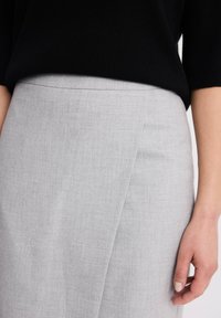Kobieta ubrana w czarny, strukturalny top włożony w jasnoszary spódnicę o kroju kopertowym, z ręką swobodnie opartą przy boku.