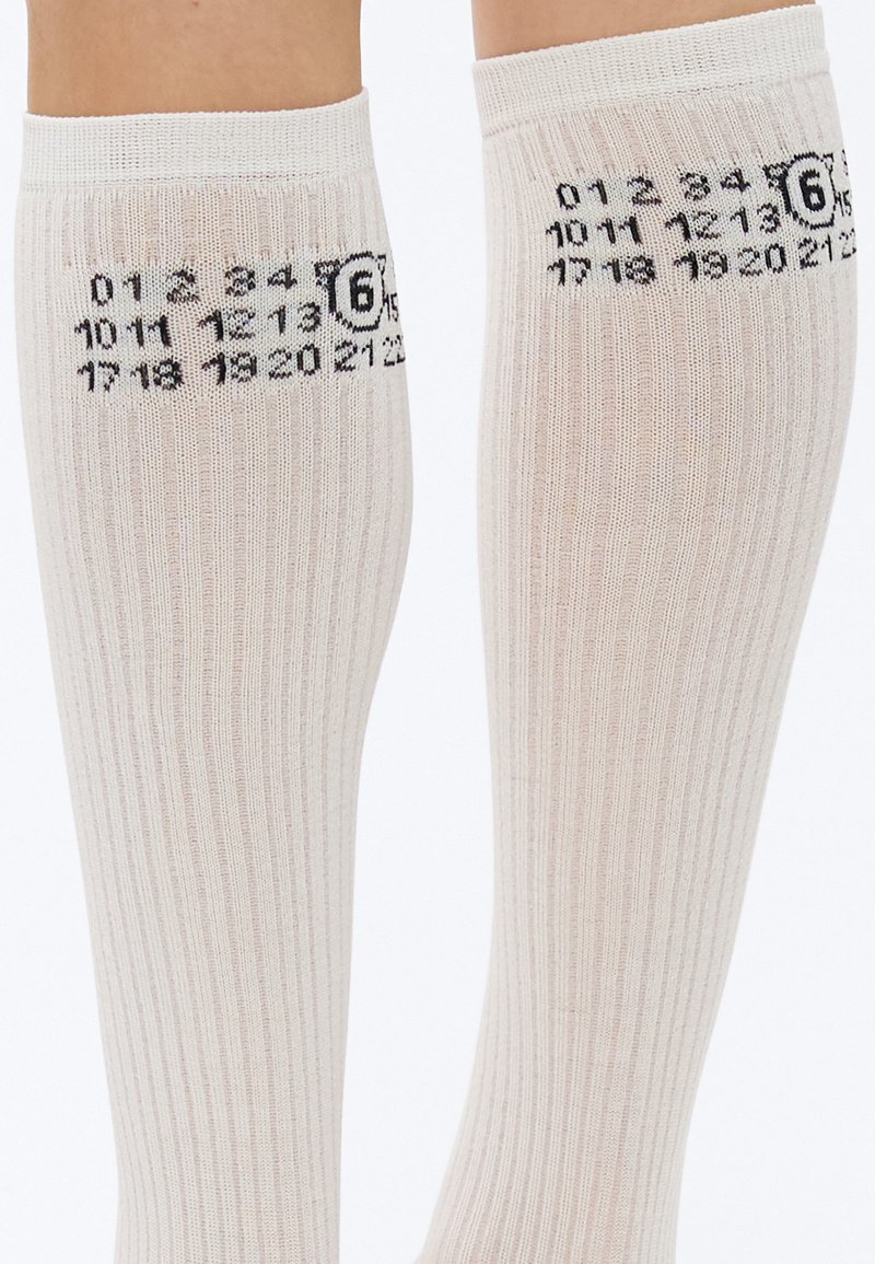 Knæhøje, ribstrikkede sokker i lys beige med sorte talmønstre langs siderne. Blød tekstur, elastisk kant øverst.