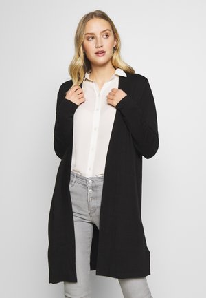 Zwarte cardigan van zacht stof, met een lange, open voorkant en aangesloten lange mouwen. G gedragen over een witte blouse met knopen.