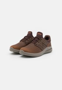 Sneaker in pelle marrone con accenti in rete, superficie testurizzata, suola grigia imbottita e cuciture a contrasto. Presenta lacci e dettagli del logo.