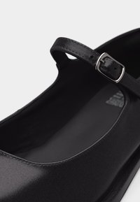 Μαύρο παπούτσι Mary Jane με ένα ρυθμιζόμενο λουράκι με αγκράφα και ματ φινίρισμα σε ανοιχτό φόντο.