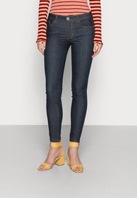 Strakke donkerblauwe spijkerbroek met een hoge taille, vijf zakken en oranje stiksels, gecombineerd met gele sandalen met blokhak.