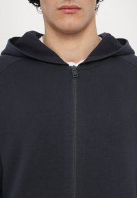 Homme portant un sweat à capuche bleu marine foncé zippé, avec une chemise blanche légèrement visible en dessous, montré du cou au haut du torse.