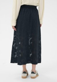 Jupe longue en tissu bleu marine transparent avec motif floral, dotée d'une ceinture élastique. Associée à des baskets décontractées, vue de dos.