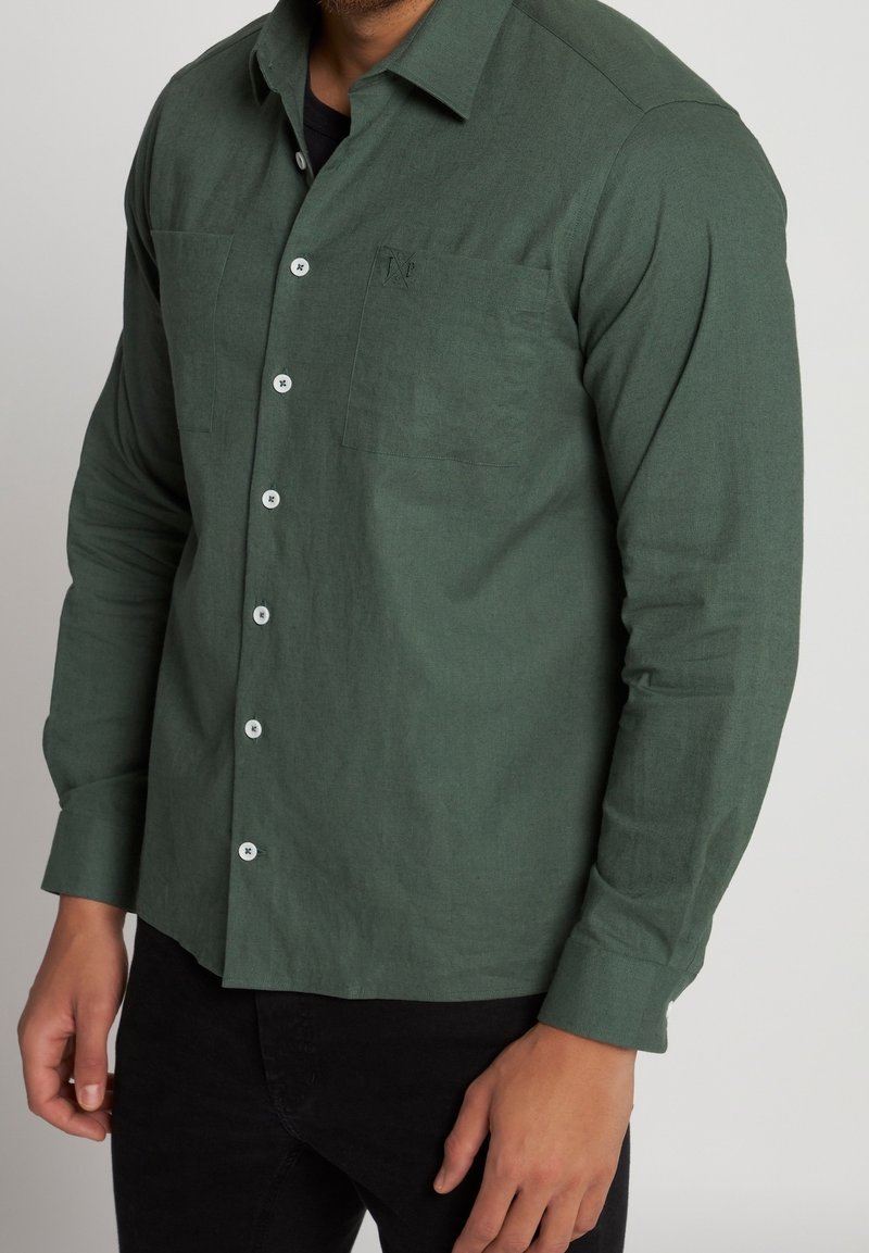 Homme portant une chemise boutonnée vert foncé avec une poche poitrine et un pantalon noir, debout devant un fond clair uni.