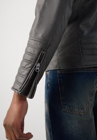 Grau Lederjacke mit gerippter Detailierung an den Ärmeln und einem Reißverschluss an den Bündchen, kombiniert mit blauen Denim-Jeans.