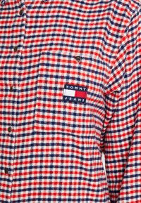 Camicia di flanella a quadri rossa, bianca e blu con bottoni, una tasca e una toppa con il logo "TOMMY JEANS" sulla parte sinistra del petto.