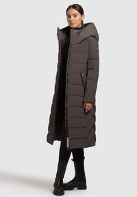 Frau in einem langen, dunkelbraunen, gepolsterten Mantel mit hohem Kragen, schwarzen Leggings und schwarzen Lederstiefeln, die vor einem schlichten Hintergrund steht.