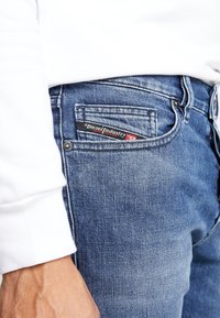 Niebieskie dżinsy z denimu o zużytej fakturze, z widoczną białą metką kieszeni z napisem „Diesel Industry”. Rękaw białej koszuli z długim rękawem jest częściowo widoczny.