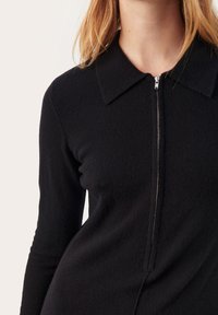 Robe noire en tricot avec un col et une fermeture éclair à l'avant. Le tissu semble doux et texturé, avec des manches longues ajustées.