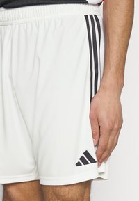 Pantalones cortos deportivos blancos hechos de un material ligero, con tres rayas verticales negras y un logo de Adidas en negro en la parte inferior izquierda.