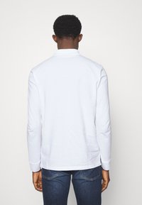 Weißes Langarm-Polohemd mit Kragen, glatter Textur und leicht abgerundetem Saum. Getragen mit dunklen Jeans.