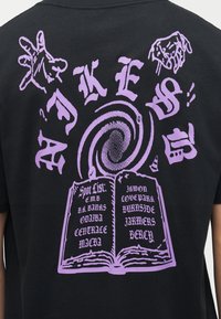 Dos d'une chemise noire avec un texte gothique violet « FAKE US » tourbillonnant au-dessus d'un livre ouvert listant des lieux comme ODAIBA et BERCY dans une police stylisée.