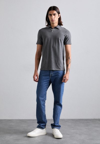 Jonge man die binnen staat in een donkergrijze polo met korte mouwen, blauwe spijkerbroeken, witte sneakers en zichtbare tatoeages op beide onderarmen.