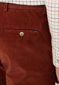 Bruine corduroybroek met een voorzak met een knoop en een merklabel. De stof heeft een gestructureerd ribbelpatroon.