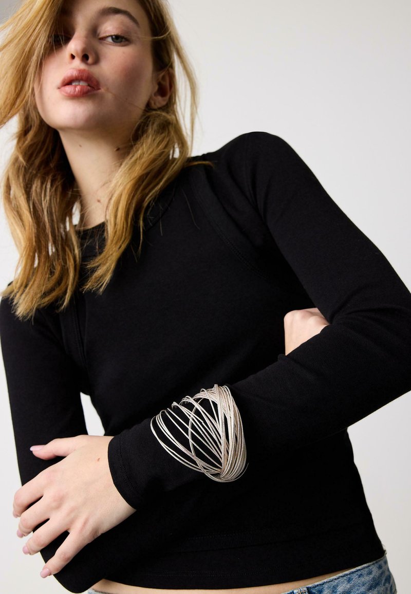 Jeune femme aux cheveux blonds portant un haut noir à manches longues et un grand bracelet en fil d'argent au poignet gauche, posant devant un fond uni.
