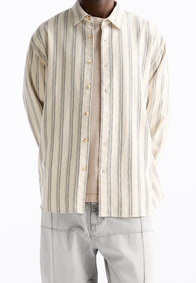 Chemise à boutons, rayée crème et gris, avec une coupe oversize, des fines rayures verticales et des boutons en bois. Portée sur un haut beige.