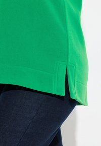 Sudadera de forro polar verde con dobladillo recto y aberturas laterales, llevada sobre unos vaqueros azul oscuro. La textura parece suave con un mínimo de detalle de costura.