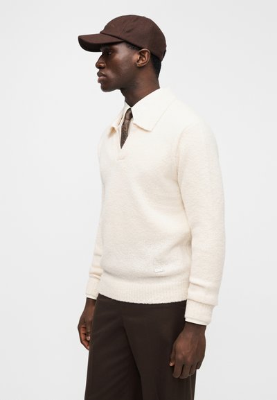 Cream-colored knitted pullover with a V-neck and wide collar. Brown cap with a curved brim and logo details. Dark brown pants.