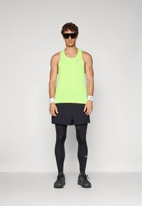 Man in bright yellow sleeveless athletic top, black running shorts over black leggings, black sneakers, white wristbands, and large black sports sunglasses.