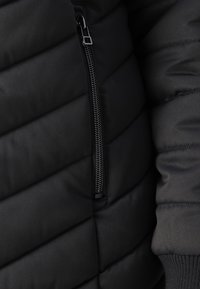Chaqueta acolchada negra con un bolsillo lateral con cremallera, que presenta una textura acolchada y puños acanalados. El material parece suave y liso.