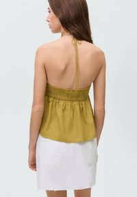 Zadný pohľad na bezrukávový olivovo zelený top so smockovaným dizajnom, skombinovaný s bielou denimovou sukňou, s nastaviteľnými ramienkami.