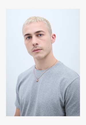 Joven con cabello rubio corto y ojos azules, vestido con una camiseta gris y un collar con un colgante dorado en forma de sol, sobre un fondo claro y liso.