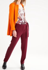 Giacca tailored arancione sopra un top con stampa geometrica multicolore, abbinata a pantaloni tapered bordeaux e stivali alla caviglia neri.