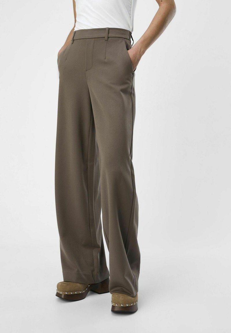 Personne portant un pantalon marron à jambes larges avec les mains dans les poches et des chaussures compensées beiges cloutées sur fond blanc.