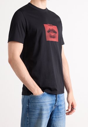 Homme portant un T-shirt noir avec un graphisme rouge de lèvres sur la poitrine et un jean bleu, main dans la poche sur fond blanc uni.