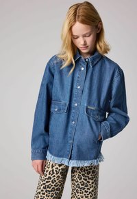 Camicia in denim di blu medio con bottoni frontali, due tasche sul petto e orlo sfilacciato. Abbinata a pantaloni con stampa leopardata in beige e nero.