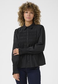Camicia nera con bottoni, dettaglio smock sul davanti, maniche lunghe con polsini smock e vestibilità morbida. Tessuto di cotone con una consistenza liscia.