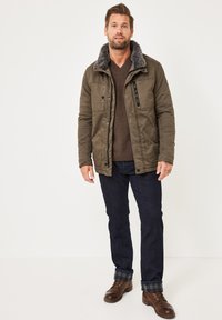 Olivgrüne isolierte Jacke mit einem grauen Fellkragen, mit Reißverschluss versehenen Brusttasche, knöpfigem Vorderteil, über einem braunen Pullover und dunklen Jeans getragen.