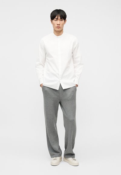 Άνδρας όρθιος, στραμμένος προς τα εμπρός, φορώντας λευκό πουκάμισο με κουμπιά, γκρι φαρδιά παντελόνια και λευκά αθλητικά παπούτσια, με φόντο ένα απλό λευκό φόντο.