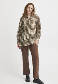 Chemise à carreaux marron avec des accents verts et cr�ème, à manches longues, boutonnière sur le devant, associée à un pantalon marron et des chaussures noires.