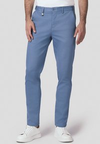 Niebieskie bawełniane spodnie chino o slim fit, z zapięciem na guzik z przodu, kieszeniami po bokach, noszone z białymi trampkami.