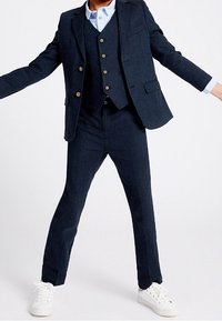 Granatowy, dopasowany garnitur trzyczęściowy wykonany z fakturowanej tkaniny, składający się z marynarki z czterema guzikami, kamizelki i slim fit spodni. Białe sneakersy.