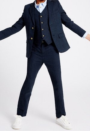 Traje de tres piezas azul marino ajustado, hecho de una tela texturizada, que incluye una chaqueta con cuatro botones, un chaleco y pantalones de corte estrecho. Zapatillas blancas.