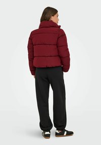 Femme en doudoune bordeaux, pantalon de jogging noir et baskets noires avec des rayures blanches, se tenant de dos face à l'appareil photo sur fond blanc.